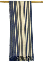 The MyHomeDecor.ca Woven Table Runner With Fringe - Blue is a long cotton woven fabric hanging on a wooden rod. The fabric features vertical stripes in various shades of blue, gray, and cream. The ends are fringed, with the fringe comprising blue and cream threads. The overall appearance is rustic and textured, making it perfect as a table runner.