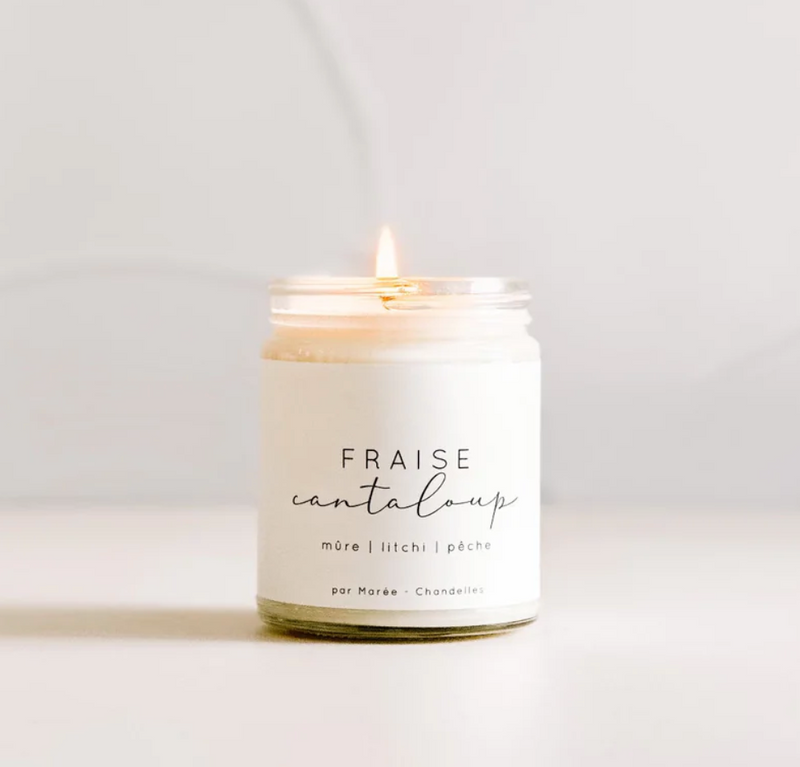 A lit soy candle in a glass jar labeled "Large Soy Candle - Strawberry & Cantaloupe" with additional text "mûre | litchi | pêche" and "par MyHomeDecor.ca." This environmentally friendly candle is placed on a light-colored surface against a light background.