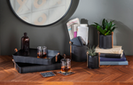 A wooden table with a herringbone pattern holding black trays, a glass of whiskey, and books next to a MyHomeDecor.ca Mini Planter with Leather Handle - Black - Small. A round mirror and a tic-tac-toe decoration hang on the wall in the background, and a small brown glass bottle adds charm.