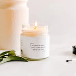 A lit Medium Soy Candle Clementine & Mango from MyHomeDecor.ca in a clear jar with a white label sits on a white surface. The label reads "un délicat merci pour votre place dans nos vies." There are green leaves to the left and a black matchstick with a white match head to the right of the candle, evoking hints of mango and clementine.