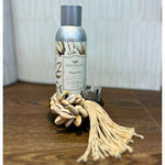 A bottle of Fall Exclusive Package Deal - 4 Pieces by MyHomeDecor.ca rests on a wooden surface. In front, a decorative shell string tassel adorned with beige seashells and white strings adds charm. Both items sit against a neutral, lightly textured background, making it the perfect host gift.