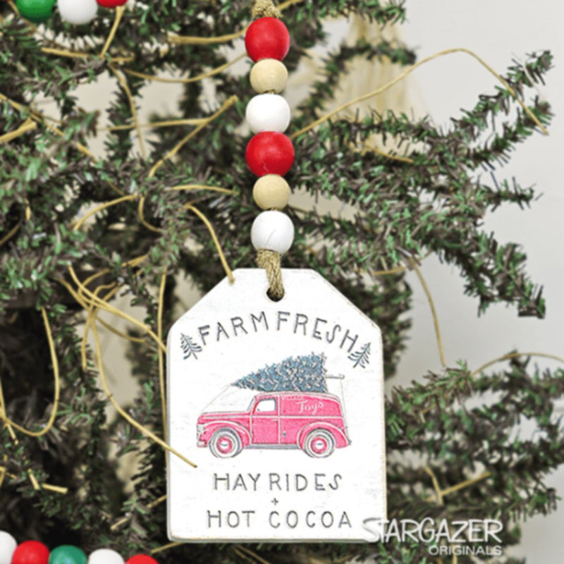 A festive Christmas ornament hangs on the tree, shaped like a truck tag with a red truck carrying a Christmas tree. It reads "FARM FRESH," "HAYRIDES," and "HOT COCOA" below it. The ornament's string is adorned with red, white, and beige beads, perfect for winter decor. This charming decoration is the Christmas Truck Tag with Beads by MyHomeDecor.ca.