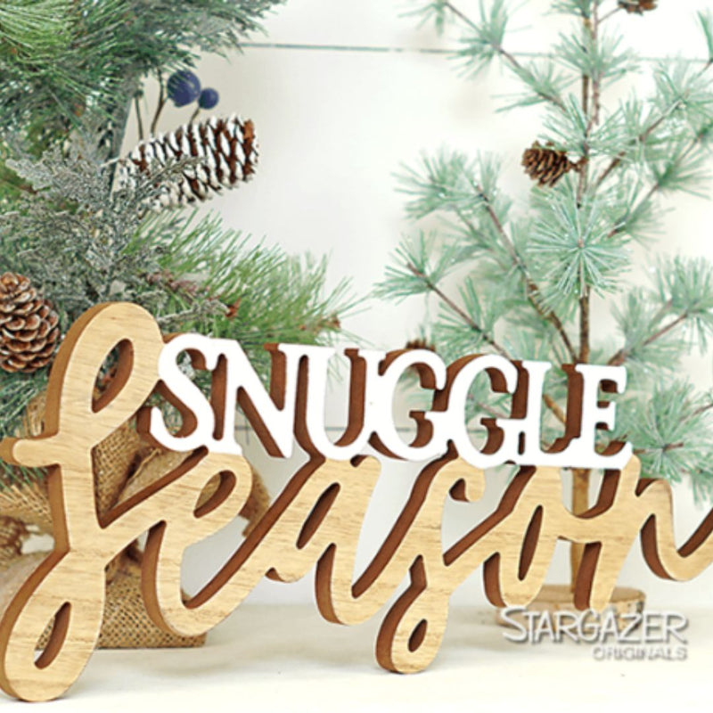 A MyHomeDecor.ca "Snuggle Season - White" decorative wood sign with the words "Snuggle Season" in a mix of white and natural wood colors is placed in front of a small pine tree adorned with pinecones. The sign sits on a light-colored surface, and the background has a festive, Christmas theme.