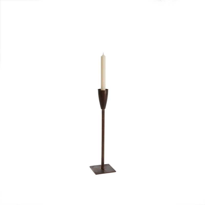 A tall, minimalist **Large El Grande Candle Stick - Leather** made of dark, rustic metal with a square base supports a single, slender white candle. Made in India, the design is simple and elegant, highlighting the vertical structure and contrasting colors of the metal and candle. This product is brought to you by **MyHomeDecor.ca**.