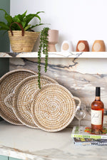 A kitchen shelf featuring woven baskets, two stacked cookbooks, a bottle of rosé wine, and two wine glasses. Potted house plants and decorative vases sit on the upper shelf, with a cute MyHomeDecor.ca Tapered Pot - Beige housing a cascading green plant hanging down. The background is a marble backsplash—perfect home decor elegance.