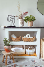 A white shelf with a dark countertop displays Medium Bright Raised Base Pots - Orange/Pink/Sandstone from MyHomeDecor.ca, wicker baskets, and decorative items. The floor features a terrazzo design. On the countertop, a black line art sculpture of a face is visible. A wooden stool with a house plant is beside the shelf.