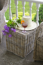 A natural straw stool on a porch is adorned with a **Woven Straw Hat - Natural** by **MyHomeDecor.ca**, a glass of wine garnished with an orange slice, and a bouquet of purple and white flowers. Surrounding the stool are wicker chairs, and the background reveals a green lawn and white railing.