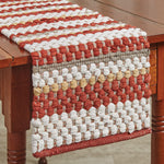 A close-up of the MyHomeDecor.ca Table Runner - Kingswood Chindi on a wooden table. The runner, perfect for farmhouse living, features horizontal stripes in shades of red, white, brown, and beige with a thick, textured weave. The wooden table has a warm, natural finish.