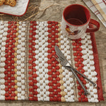 A rustic table setting features a red and white woven placemat, a red mug with a rooster design, and a fork and knife crossed on the placemat. The background includes a wooden surface with light-colored straw-like materials scattered around, perfect for farmhouse living with the Table Runner - Kingswood Chindi from MyHomeDecor.ca.