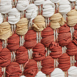 Close-up of a textured woven artwork, reminiscent of a MyHomeDecor.ca Table Runner - Kingswood Chindi, featuring rows of tightly packed, small, spherical fabric pieces in shades of white, beige, and red-orange. The fabric shapes are layered and intricately interlaced with vertical strands, creating a tactile surface perfect for farmhouse living.