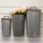Three wall-mounted Large Galvanized Wall Vases - Grey from MyHomeDecor.ca are displayed against a white wooden wall, evoking a sense of nostalgia. The vases vary in size, with the smallest on the left and the largest on the right. Classic forms shine through, especially in the middle vase that holds light pink and white flowers.