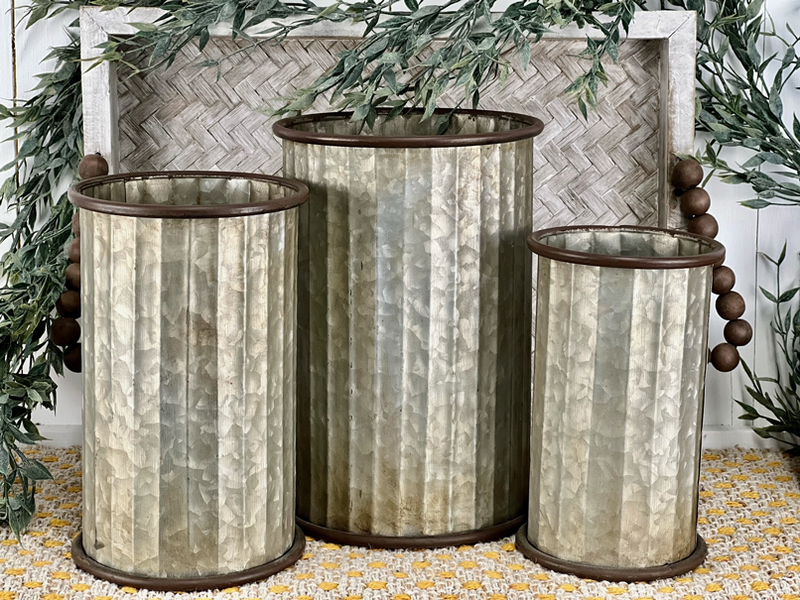 Three rustic MyHomeDecor.ca Large Bucket - Galvanized planters of different sizes sit on a woven yellow and white rug, perfect for your spring decor. They're placed in front of a rectangular wicker tray. Greenery and wooden beads drape around the scene, adding a touch of natural home decor charm.