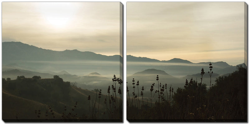 A serene landscape of rolling hills shrouded in mist, with soft sunlight filtering through a cloudy sky. Silhouetted wildflowers and tall grasses in the foreground, distant mountains enveloped in fog fading into the horizon under a calm, hazy atmosphere. Perfect as MyHomeDecor.ca Canvas - Fog in the mountains for an elegant set of 2.