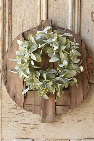 A Candle Ring – Grand Bay from MyHomeDecor.ca, reminiscent of Spring Decor, hangs on a rustic, circular wooden board with a cross pattern. The background consists of a weathered wooden door with visible texture and vertical ridges.