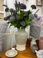 A large Keegan Vase - Two Tone from MyHomeDecor.ca filled with purple and lavender flowers sits on a wooden surface. Surrounding the vase are various decorative items, including a bird-shaped jewelry holder, smaller blue-green vases, and fabric quilts with intricate patterns in the background.