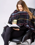 A person with long hair, wearing a black shirt and black pants, sits in a black leather chair, covered with a Deluxe Knee Blanket - Spirit of The Titanic by MyHomeDecor.ca. They are holding an open book in one hand and a mug with a green and yellow design in the other, appearing to read attentively.
