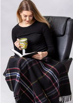 A woman with long hair sits in a black armchair, wearing a black long-sleeve top and a MyHomeDecor.ca Deluxe Knee Blanket - Spirit of The Titanic on her lap. She is reading a book and holding a yellow patterned mug. The background is plain white.