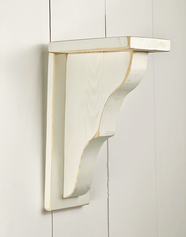 A Plain Corbel – White mounted on a white wall. The corbel has a decorative curved design and is painted in a light cream color. The edges of the bracket are slightly distressed, giving it a rustic look—a perfect touch for spring decor or any home decor style from MyHomeDecor.ca.